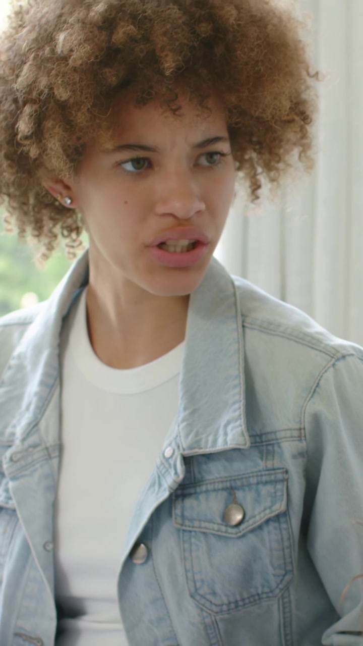 Vertical video: Lifting head after pausing, woman in denim jacket shifting gaze, frowning at home