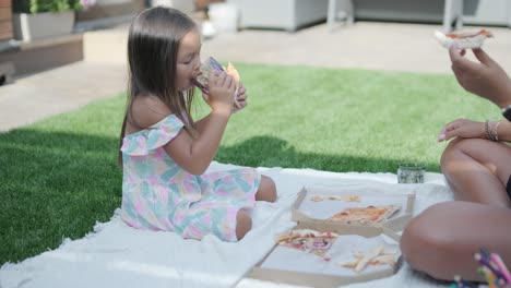 a mother and daughter enjoy a summer picnic outdoors on a grassy lawn, sharing pizza and spending quality time together on a sunny day. the family bonding scene reflects leisure and relaxation.