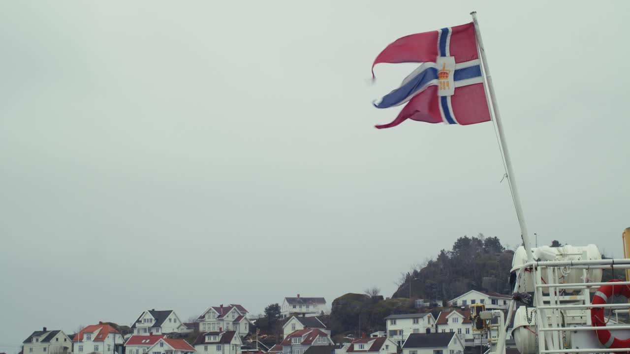 Et statisk etableringsbilde av Norges treflikede flagg, med svalehaledesign, og bolighus i Kragerø i Sørøst-Norge på en overskyet dag.