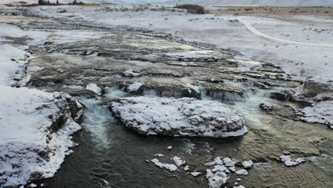 Luftaufnahme-Stromaufwärts-Entlang-Eines-Flusses-In-Island,-Vorbei-An-Einer-Winterlichen-Berglandschaft-Win