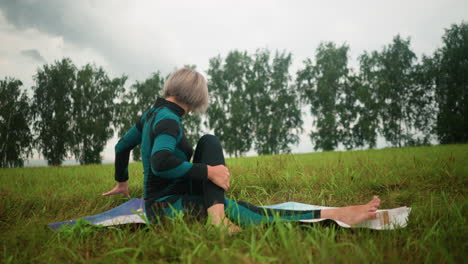 old woman seated on yoga mat practicing seated twist yoga, turning to the left with head slightly lifted backward, in a vast grassy field with trees lined up in the distance under a cloudy sky