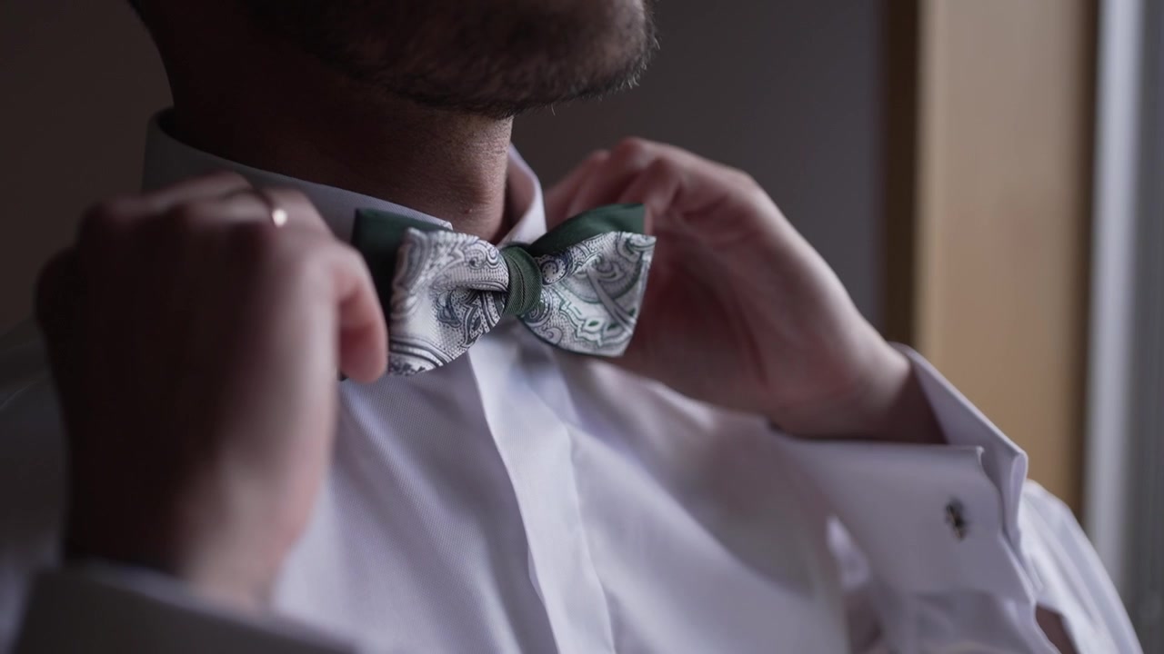 Uomo che si sistema il papillon a fantasia, con gemelli e fede nuziale, in primo piano.
