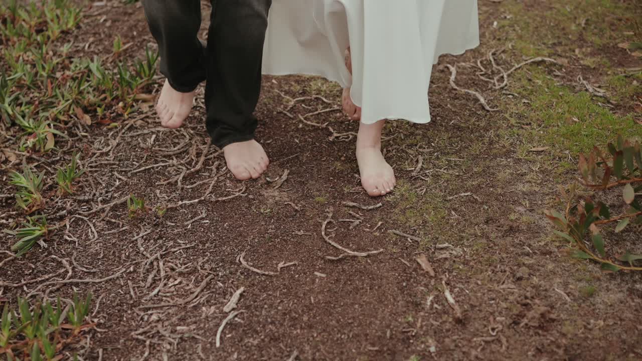 Lower body view of couple walking barefoot on soil path with roots while holding hands in natural outdoor setting