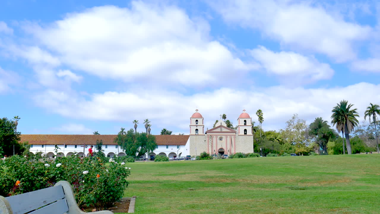 푸른 하늘 아래 산타 바바라 미션 빌딩은 캘리포니아 잔디밭 건너편 장미 정원의 빈 벤치에서 바라볼 수 있습니다.