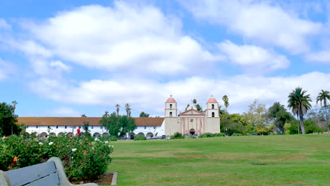 Das-Missionsgebäude-Von-Santa-Barbara-Unter-Blauem-Und-Bewölktem-Himmel,-Betrachtet-Von-Einer-Leeren-Bank-Im-Rosengarten-Auf-Der-Anderen-Seite-Der-Wiese-In-Kalifornien