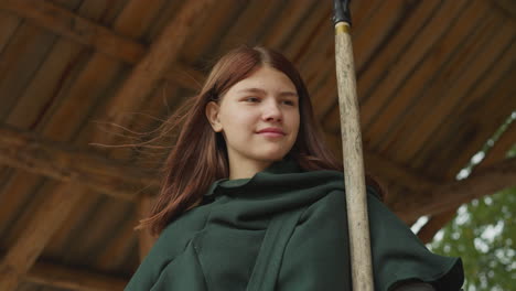 smiling girl in green cloak stands in gazebo holding spear