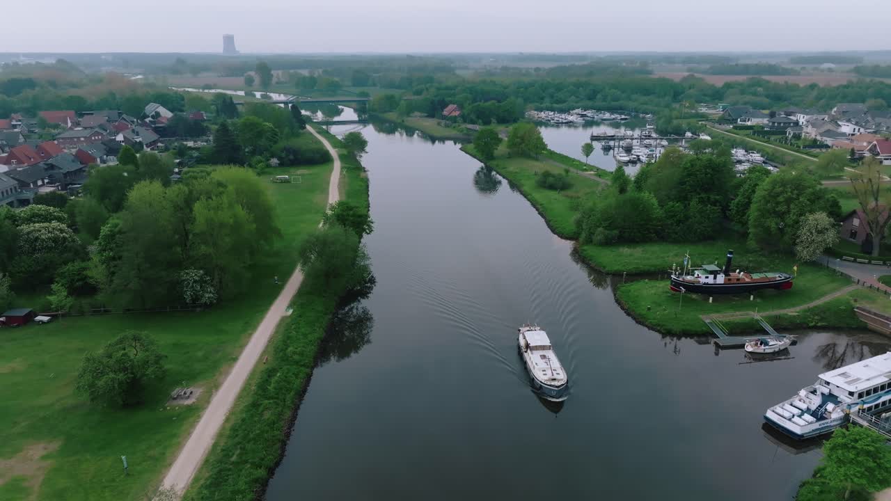 Aerial zoom out as a barge approaches a marina in northern Germany. Surrounded by green banks and calm waters on a cloudy morning.