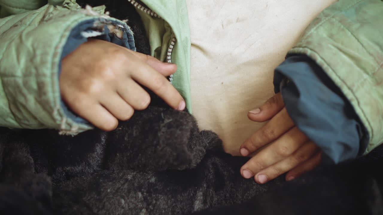 Close up of dirty hands of young person resting on worn blanket, wearing old green jacket and faded shirt, evoking sense of poverty, cold, fatigue, and vulnerability in harsh living conditions