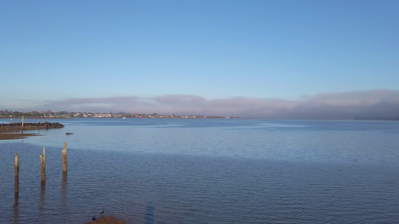 A pan across harbour water with wooden posts in foreground