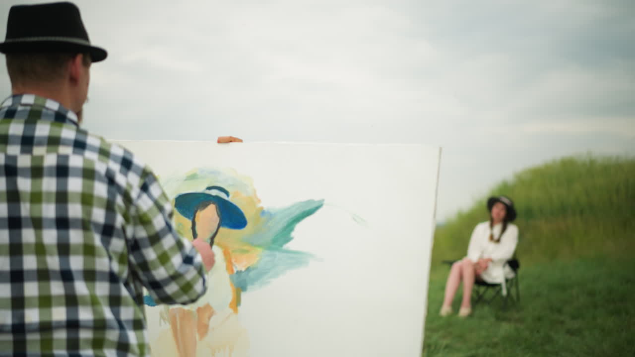 An artist, wearing a hat and a checkered shirt, is focused on painting a portrait on a large canvas while holding a palette. In the background, a blurred woman is seated on a chair in a grass field