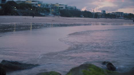 el sol poniente derrama colores pastel de rosa, azul y naranja en el cielo, las nubes y el agua son empujadas suavemente a la orilla de una playa de arena limpia mientras los transeúntes disfrutan de su tarde fresca