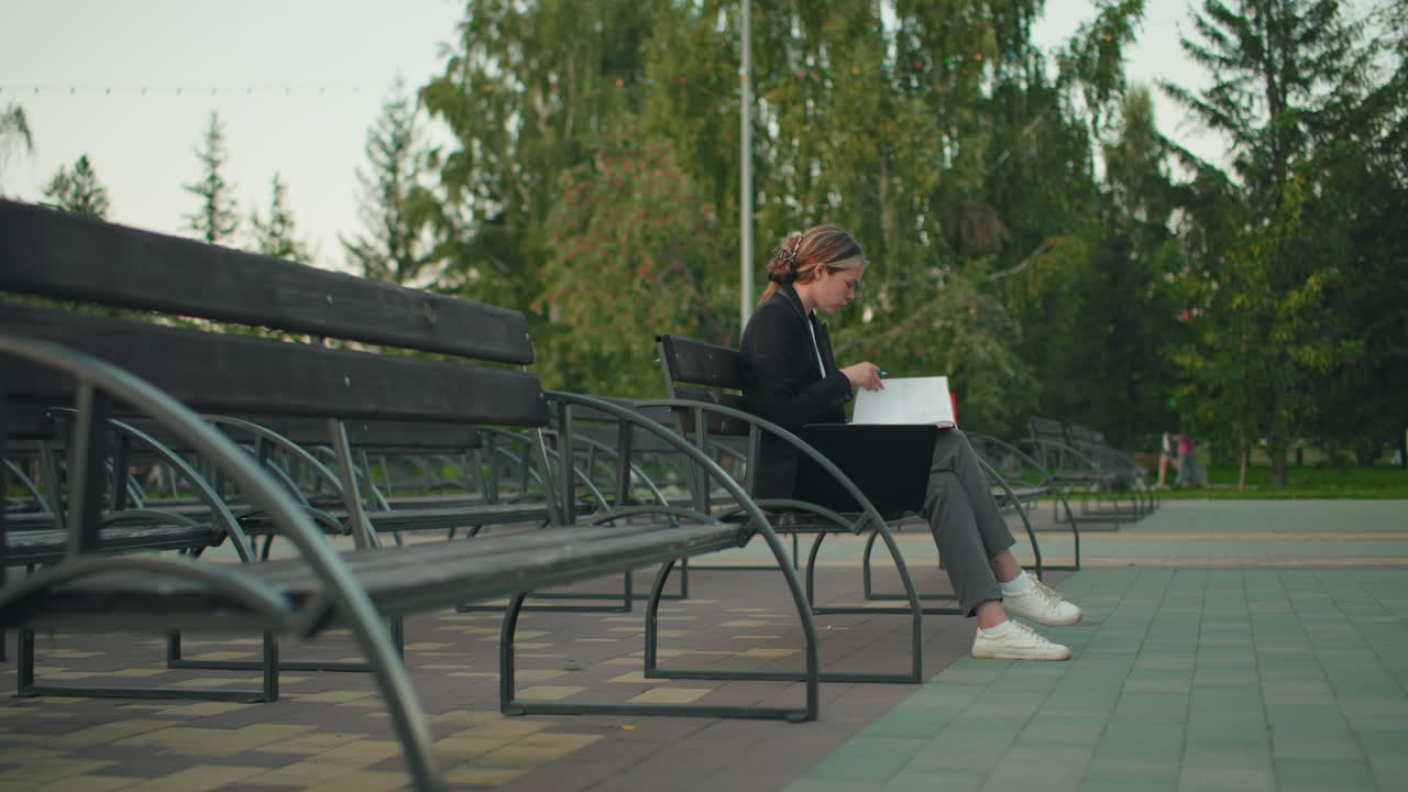 Young woman in professional attire seated alone on outdoor bench flipping through red folder with pen, laptop beside her, in calm park surrounded by trees, benches, and distant people in blur view