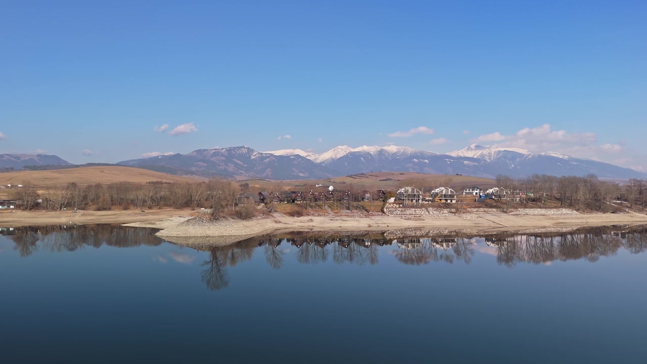 Slovakya'daki Liptovský Trnovec ve Liptovská Mara rezervuarı, karlı Yüksek Tatralar, sakin su, kıyıdaki kulübeler ve erken ilkbaharda çıplak ağaçlarla, drone ile çekilmiş