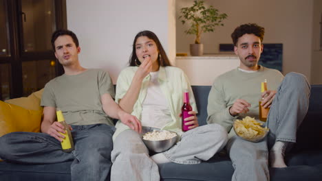 three friends watching interesting movie on television sitting on couch, eating popcorn and chips and drinking soda 1