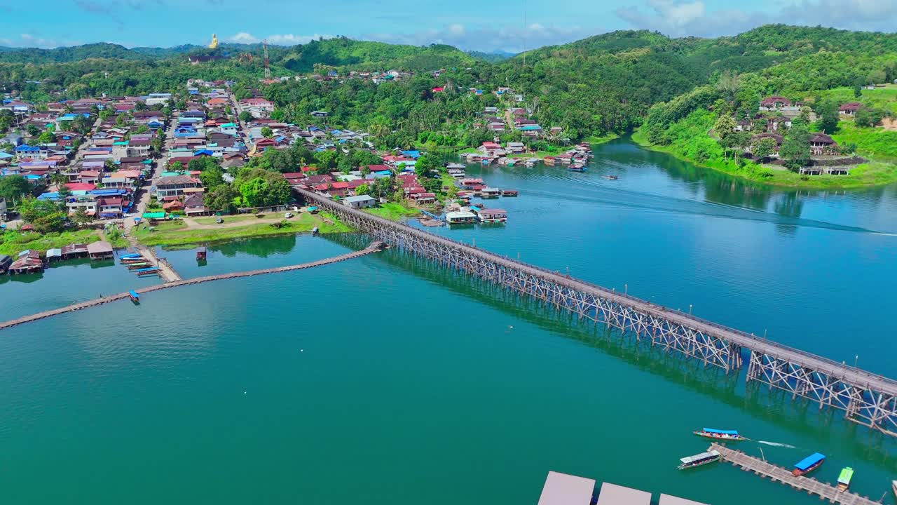 Aerial orbit of the Mon Bridge in Sangkhlaburi, Thailand. Wide view captures the wooden structure, floating houses, blue lake, village, surrounding green mountains, temples, and giant Buddha statue