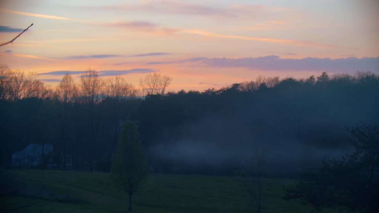 una hermosa y vívida puesta de sol de principios de primavera en el campo, con una casa que libera humo en el fondo y árboles en el medio y en primer plano