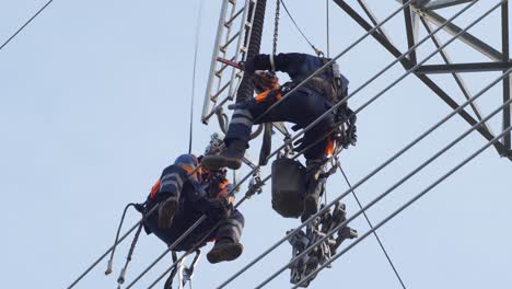 two utility workers in hardhats sit on electrical high voltage power lines for repair