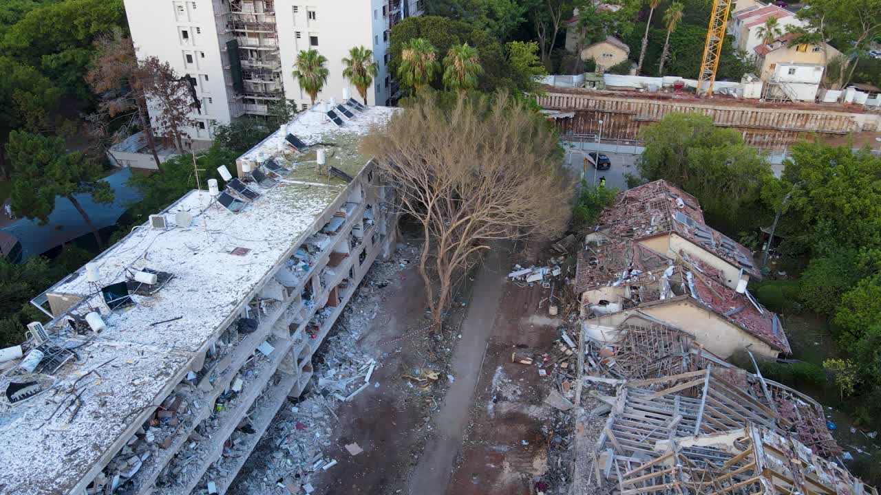 Drone circles a tree damaged by an Iranian strike in Tel Aviv, surrounded by two heavily destroyed buildings and widespread debris.