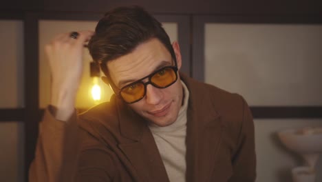 stylish caucasian man in aviator sunglasses smiles while fixing hair in the house