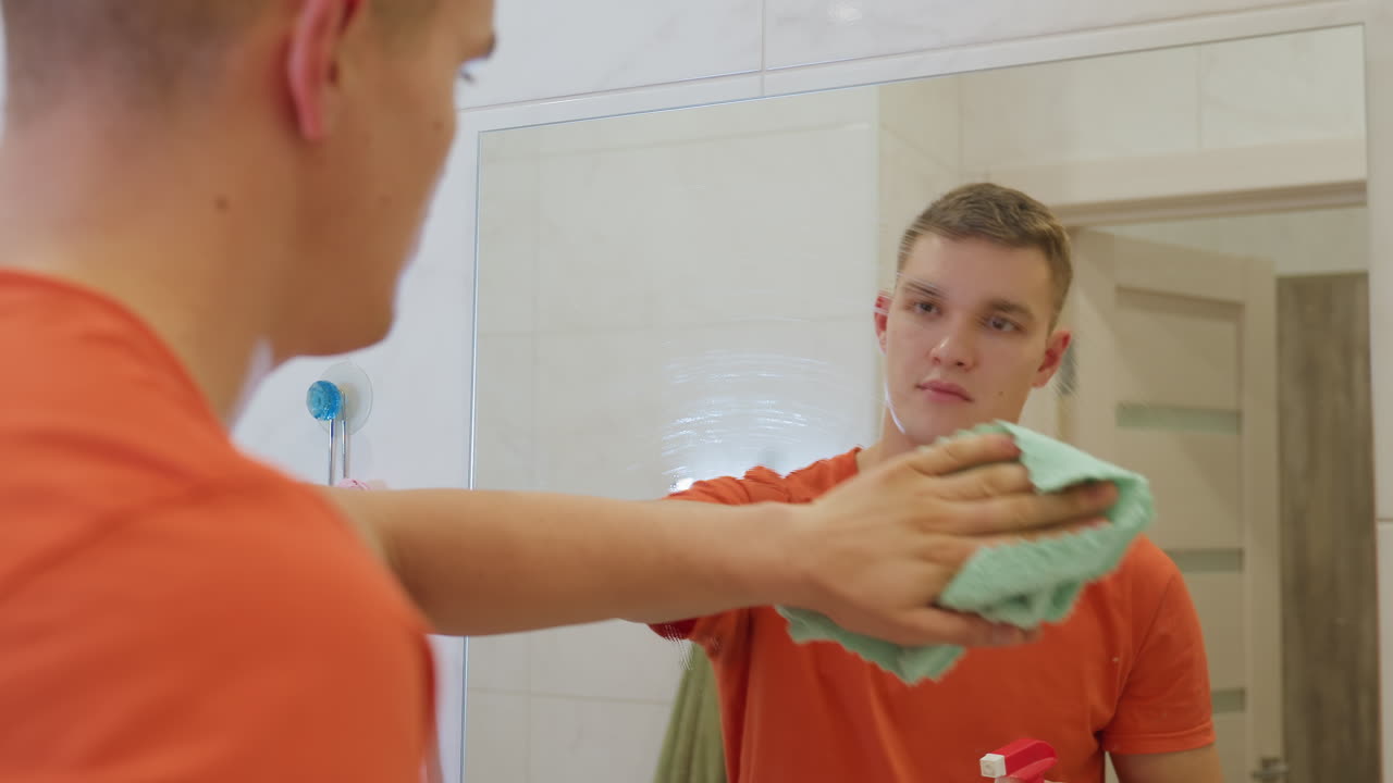 Man in orange shirt spraying soapy water onto bathroom mirror and preparing to wipe surface with soft rag, showing attention to cleanliness, household hygiene and daily routine care