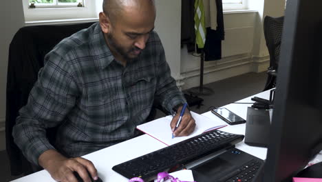 Shot of a brown man working at his office and jotting down some notes from his computer