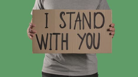 man holds in front of him a poster from a cardboard box with the words i stand with you. protest against police brutality terror and racism. isolated a green screen, chroma key. close up. slow motion