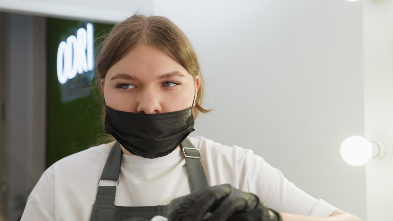 Beautician wearing black mask and apron prepares cotton pad for cleaning client eyebrow, focused on task in well-lit salon setting with soft background and beauty equipment in view