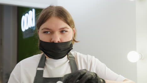 beautician wearing black mask and apron prepares cotton pad for cleaning client eyebrow, focused on task in well-lit salon setting with soft background and beauty equipment in view