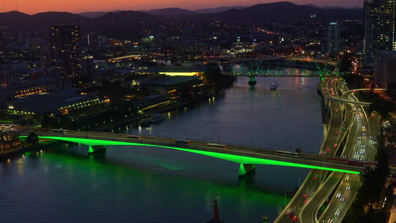 Dusk to night time-lapse shot capturing the vibrant Brisbane city, busy riverside motorway traffics, buses crossing on illuminated Victoria bridge and ferries cruising on the river.