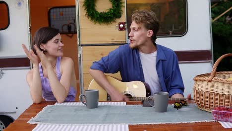 positive couple sitting at the picnic table outdoors in front their home trailer and man playing the guitar and sings song actively