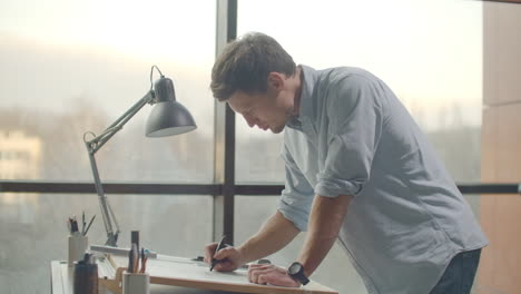 Architect-man-sits-at-drafting-table-in-modern-industrial-office-during-the-day.-Architect-working-on-blueprint-with-spesial-tools-and-pencil-close-up.-Male-architect-at-work-only-hands-with-ruler-and-pencil.-Design-of-the-house.-Engineer.-Tools-for-drawing.-Creating-a-drawing.