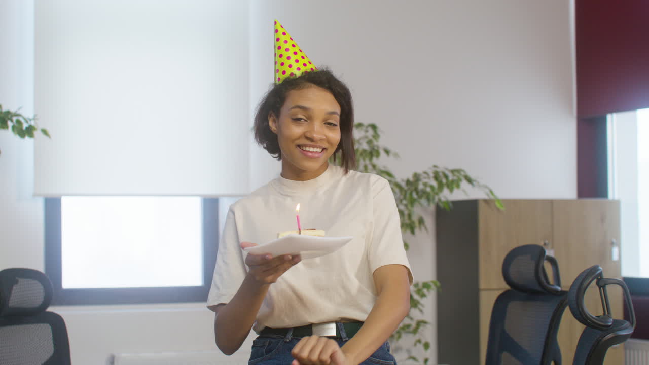 retrato de una chica estadounidense feliz sosteniendo un trozo de pastel de cumpleaños en un plato mientras mira la cámara en la fiesta de la oficina