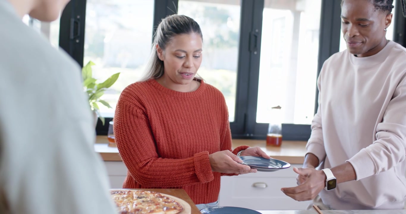 Friends slicing fresh round pizza on wood board with cutter at kitchen counter, placing dark plates