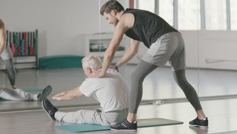 Sport-coach-helping-retired-man-doing-stretch-exercise-in-fitness-club.