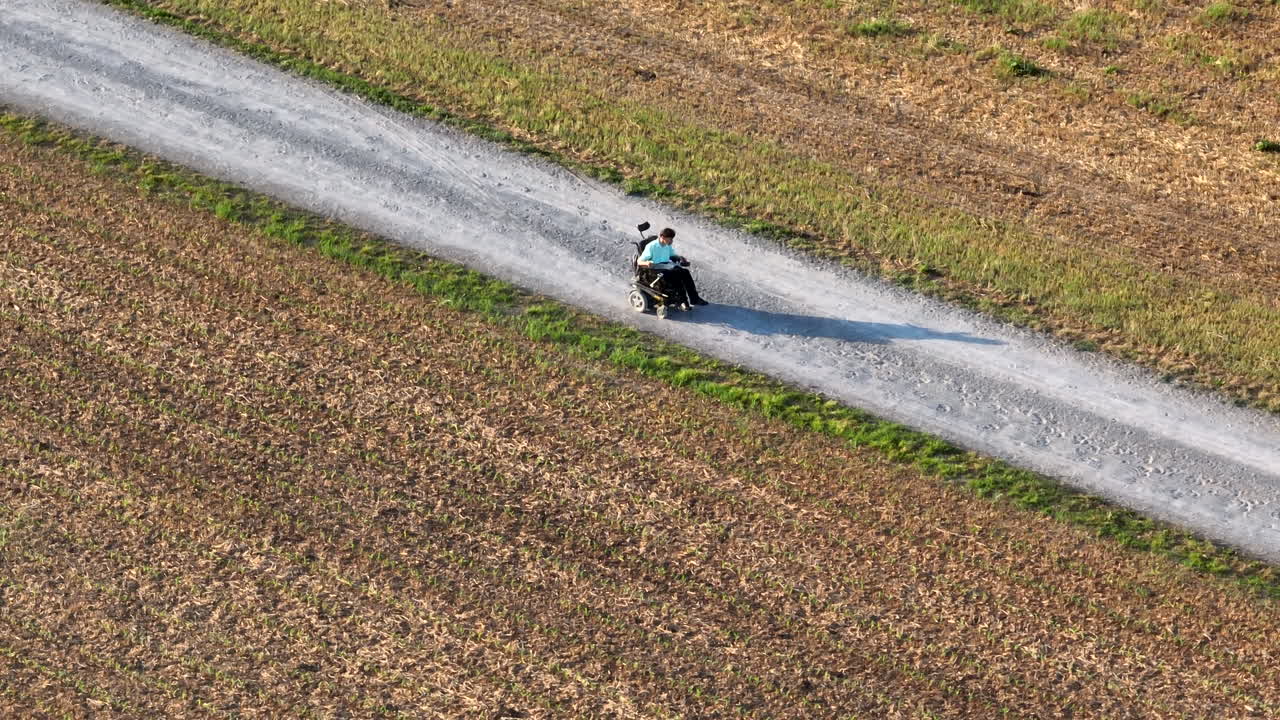 Disabled Amish man in motorized wheel chair. Long shadow as young man drives down gravel road