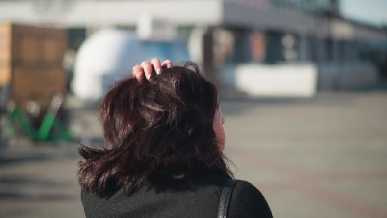 검은 코트를 입은 갈색 머리카락의 여자가 그녀의 머리카락을 조정하고, 왼손에 색 은 손을 과시하며, 빛 도시 거리에서 배경에 건물 흐릿함이 있습니다.