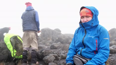 a white girl sits and rests in a jacket on a stone block during the fog while hiking through the wasteland
