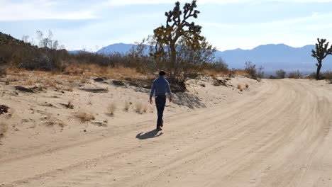 A-man-walking-alone-in-slow-motion-down-a-hot-dry-desert-road-with-Joshua-trees-and-mountains-in-the-landscape