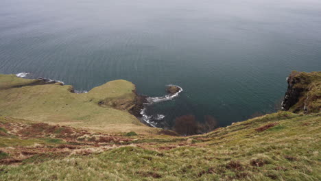 Panning-shot-from-the-edge-of-a-cliff-of-Atlantic-Ocean-waves-splashing-the-coast-on-a-rainy-and-cloudy-day-in-Scotland,-Isle-of-Skye