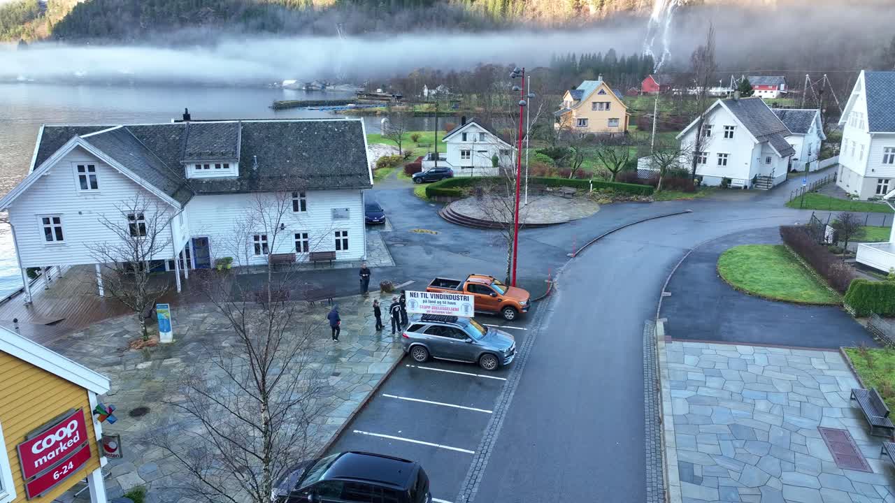Drone circles and reverses from environmental protest car in Modalen town center, revealing fjord, mountains, and tourist catamaran with scenic fog