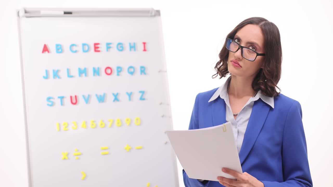 A woman, possibly a teacher or tutor, stands next to a whiteboard displaying the alphabet and numbers, while holding documents