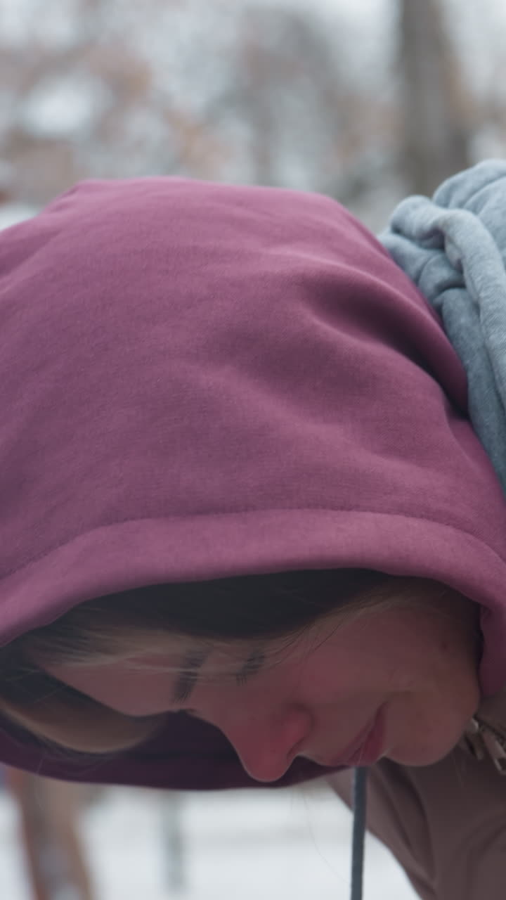 겨울 옷을 입은 젊은 여자가 추운 느낌으로 앞으로 구부러져 눈이 흐린 배경과 벌거벗은 나무, 후드 재이 머리를 고, 추운 날씨는 야외 겨울 환경에서 볼 수 있습니다.