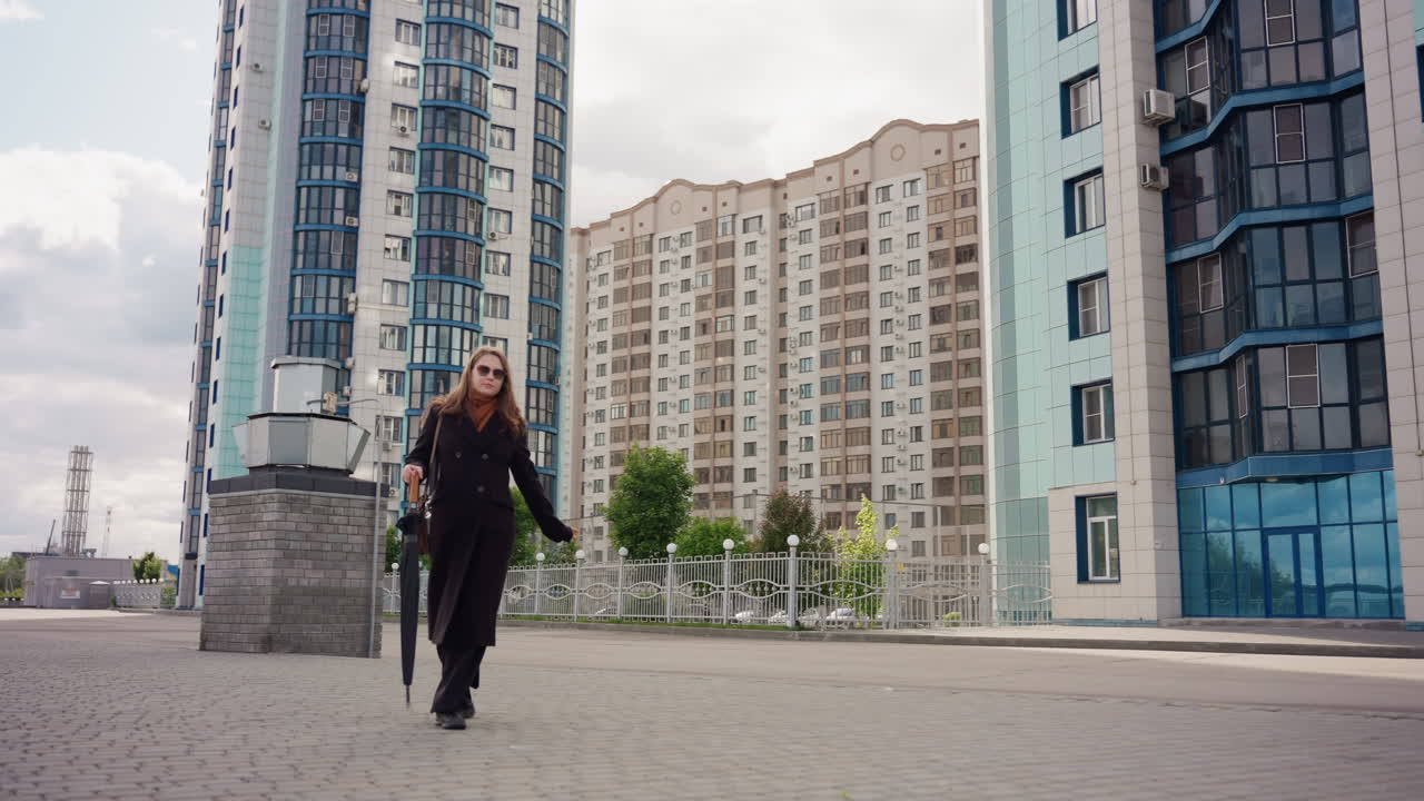 Beautiful capturing of residential tower complex radiating from background as charming woman walks across urban plaza with folded umbrella, long coat, ground level wide view under moody sky
