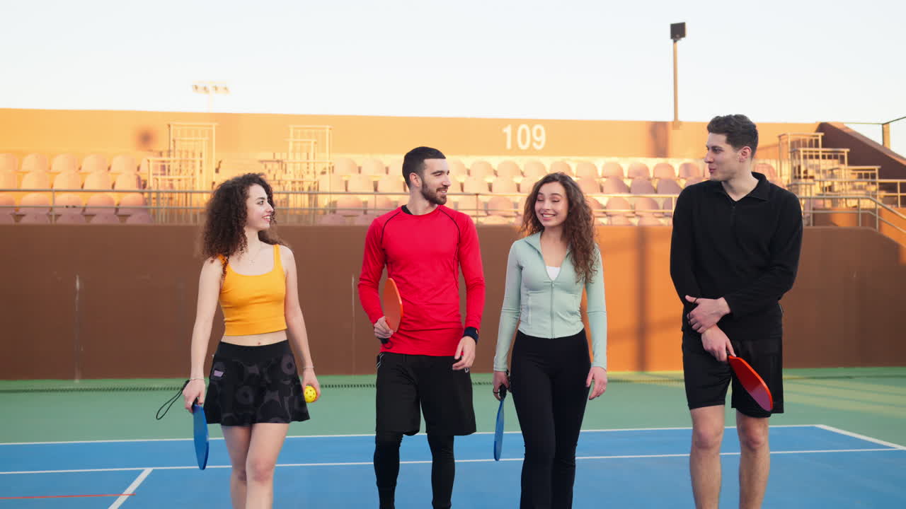 Two men and two women holding pickleball rackets walking, talking and smiling on a court. Group of friends