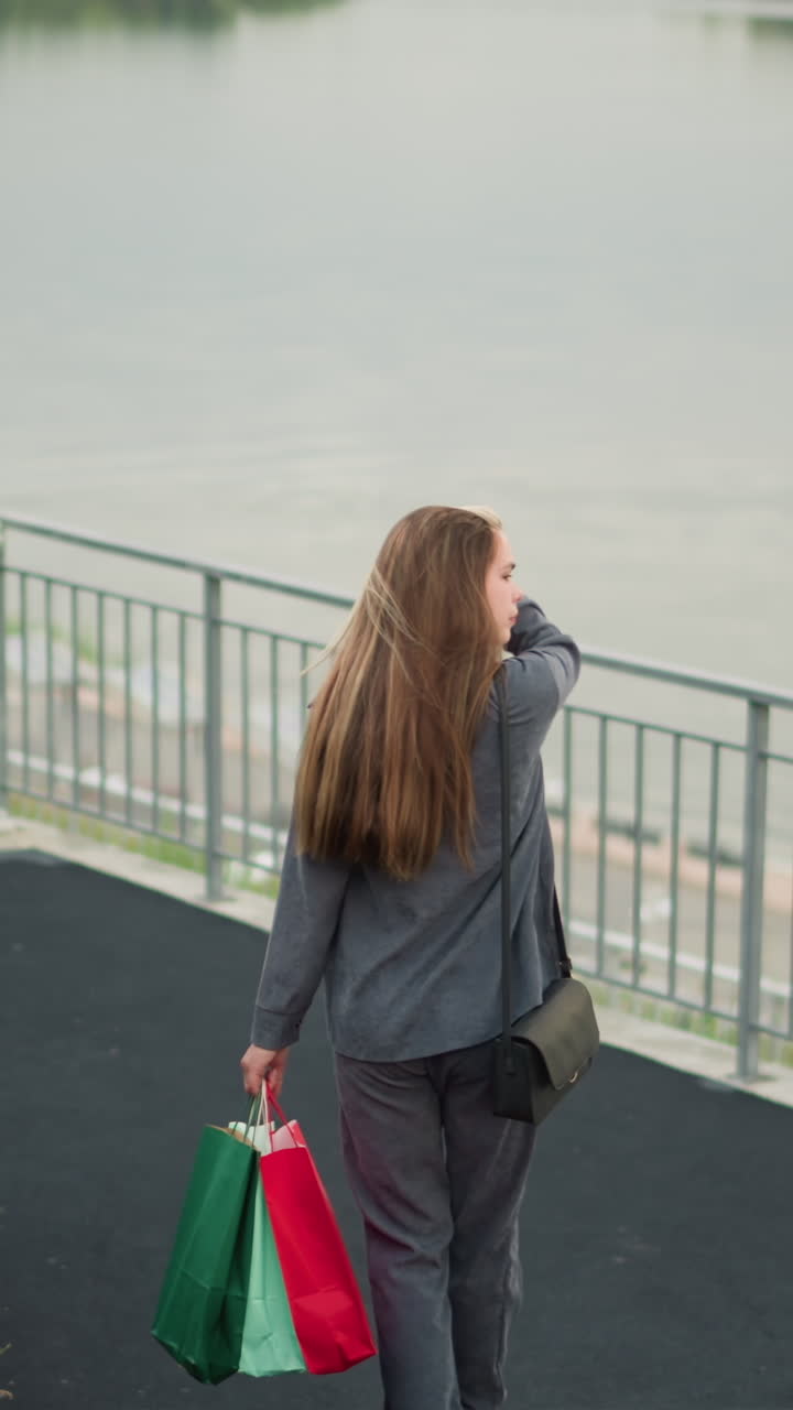 vista trasera de una mujer que lleva bolsas de compras coloridas caminando por las escaleras mientras ajusta su cabello, girando ligeramente a la izquierda cerca de la parte inferior de las escaleras, el fondo presenta un río tranquilo, un poste de lámpara y vegetación