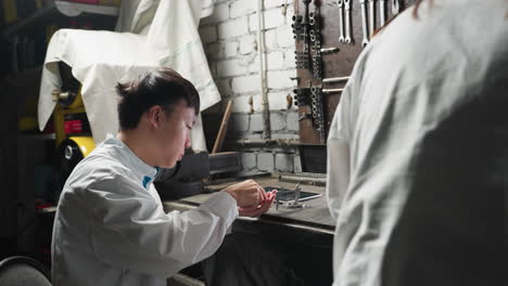 close-up back view of person observing as young technician in white lab coat works on tablet and picks up precision equipment in automotive workshop with tools and machinery in background