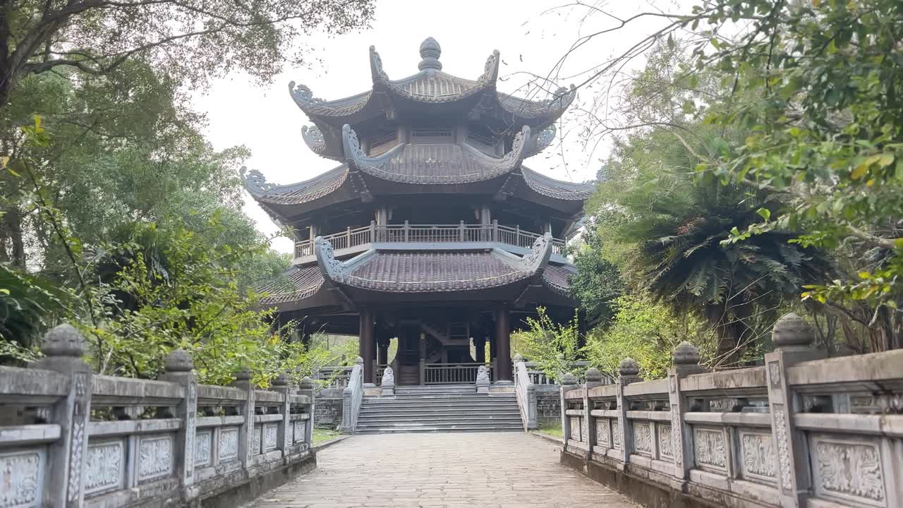 Traditionelt pagodetempel ved Bai Dinh-komplekset i Ninh Binh, set fra en stenbro og gangbro, omgivet af træer og grønt i fredfyldte kulturelle omgivelser