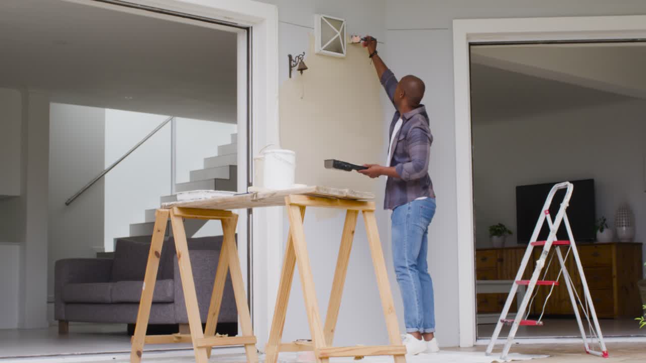 Uomo afroamericano che usa un pennello da un vassoio su un cavalletto di supporto, dipingendo la parete del portico vicino a una bocchetta di ventilazione, con spazio per il testo.
