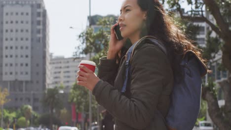 Mujer-Asiática-Sosteniendo-Una-Taza-De-Café-Hablando-Por-Teléfono-Inteligente-Mientras-Está-De-Pie-En-La-Calle