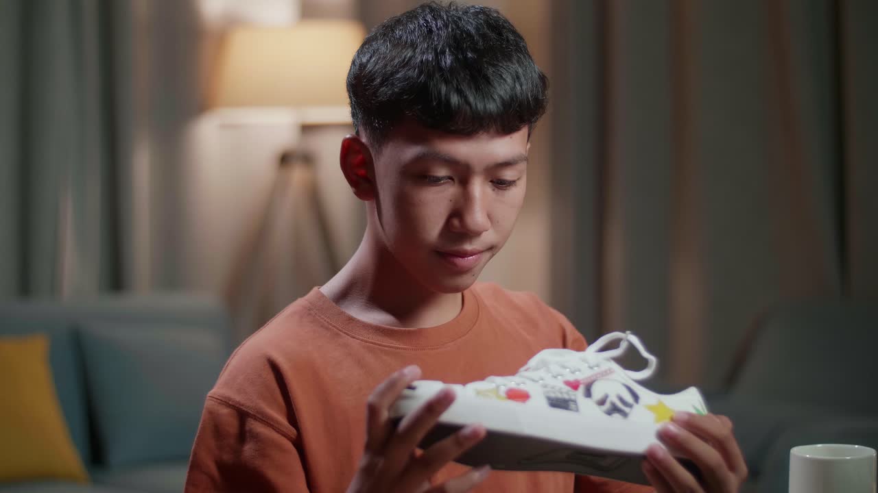 미소 짓는 아시아 소년 신발 디자이너가 다채로운 패턴의 스니커를 들고 집에서 데스크 컴퓨터에서 작업하는 동안 카메라에 엄지손가락을 보여주고 있습니다.